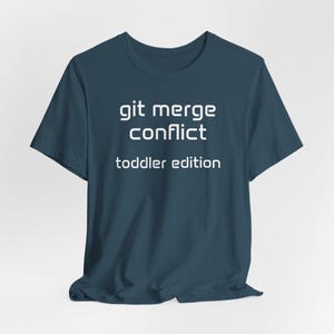 May include: A dark teal t-shirt with the text "git merge conflict toddler edition" printed in white. The t-shirt has a crew neck and is likely made from a soft fabric.