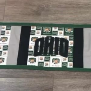 Puede incluir: Un camino de mesa rectangular con la palabra "OHIO" en letras negras. El camino de mesa tiene un fondo gris con rayas negras y blancas y diseños en forma de corazón verdes. Los bordes están ribeteados con tela verde.