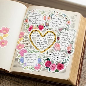 Puede incluir: Una colección de pegatinas florales con versículos bíblicos, incluyendo una pegatina en forma de corazón con las palabras «PAZ» y «Juan 14:27». Las pegatinas presentan varios arreglos florales en rosa, rojo y amarillo, sobre el fondo de un libro abierto.
