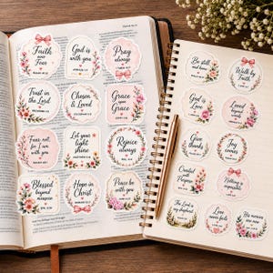 Puede incluir: Una colección de pegatinas decorativas con motivos florales y textos religiosos, dispuestas en las páginas de un libro abierto y un cuaderno de espiral. Las pegatinas incluyen frases como Faith over Fear y God is with you.