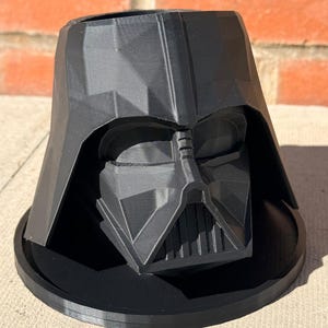 Puede incluir: Una maceta negra impresa en 3D con la forma del casco de Darth Vader. La maceta tiene una base ancha y una representación detallada de la máscara del personaje. La maceta está hecha de un material negro mate y se encuentra sobre una superficie de hormigón.