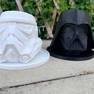 Puede incluir: Dos macetas decorativas con forma de personajes de Star Wars. Una blanca con forma de casco de Stormtrooper y otra negra con forma de casco de Darth Vader. Ambas están sobre bases redondas, colocadas sobre una superficie de hormigón.