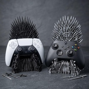 May include: Two video game controller stands shaped like thrones made of swords. One stand is black and holds a white controller, while the other is silver and holds a black controller. Several small sword replicas lie on the surface nearby.