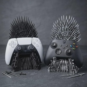 May include: Two gaming controller stands shaped like thrones made of swords. One stand is matte black holding a white and black controller, and the other is metallic silver holding a black controller with coloured buttons. Small sword replicas lie nearby.