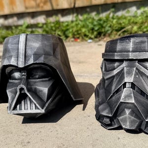 Peut inclure: Deux pots de fleurs imprimés en 3D en forme de casques géométriques. L'un est un casque de Dark Vador noir et l'autre un casque de Stormtrooper noir. Les deux présentent une finition brossée argentée métallique sur les surfaces facettées.