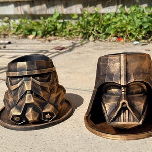 Peut inclure: Deux jardinières géométriques en forme de casques de personnages de science-fiction, avec une finition patinée bronze métallique et noir. Chaque casque repose sur une base circulaire assortie, présentant des surfaces angulaires et facettées.