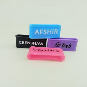 May include: Four colorful silicone wristbands. The wristbands are blue, black, purple, and pink. Each band has white text: "AFSHIR", "CRENSHAW", "Deb", and "Cynthia G.". The pink band has a heart symbol.