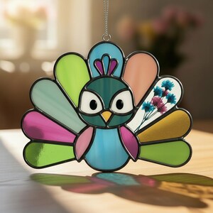 May include: A stained glass peacock decoration with a hanging chain. The peacock has a teal body, blue chest, and a fan of colourful feathers in shades of pink, green, and orange. The ornament includes a section with dried flowers.