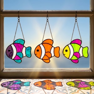 May include: Three stained glass fish in vibrant colours hang from chains in front of a window. The fish are in shades of purple, orange, yellow, green, and blue. The window frame is brown, and the background is a bright blue sky.