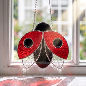 May include: A stained glass ladybug suncatcher featuring textured red wings with black circular spots, a black head, and a clear textured lower body section. The piece is suspended by a silver metal chain and displays vibrant light refraction.