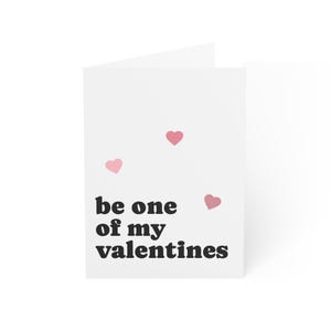 May include: A white Valentine's Day card with the text "be one of my valentines" in black. Three pink hearts are scattered across the card. The card is folded and standing upright.