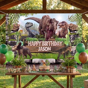 May include: A large rectangular backdrop featuring a prehistoric jungle scene with multiple dinosaurs and the text "HAPPY BIRTHDAY JASON" on a wooden sign. In the foreground, a round cake with dinosaur toppers sits on a wooden table.