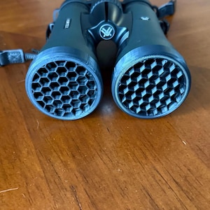 May include: Black Vortex binoculars featuring custom 3D-printed honeycomb anti-reflective lens covers. The device includes a central focus wheel, a Vortex logo on the bridge, and black adjustable straps attached to the sides of the optical housing.