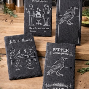 Könnte beinhalten: Eine Sammlung gravierter Schiefer-Servierbretter. Ein Brett zeigt ein Paar mit dem Text "George & Ina - Perfect Match". Ein anderes zeigt einen Raben und die Worte "Pepper" und "Salt". Das andere Brett zeigt ein Paar mit dem Text "Julia & Thomas".