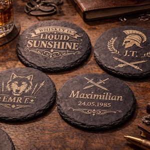 Peut inclure: Ensemble de quatre dessous de verre ronds en ardoise gris foncé avec des motifs gravés. L'un porte l'inscription "Whiskey is Liquid Sunshine", un autre présente un casque romain, et deux autres ont des textes et graphiques personnalisés. Les dessous de verre sont sur une surface en bois.