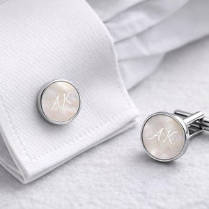 Könnte beinhalten: Silberne Manschettenknöpfe mit Perlmutt-Oberfläche, die die Initialen "AK" in Schreibschrift zeigen. Die runden Manschettenknöpfe sind auf einem weißen Hemd abgebildet, ein klassisches Accessoire.