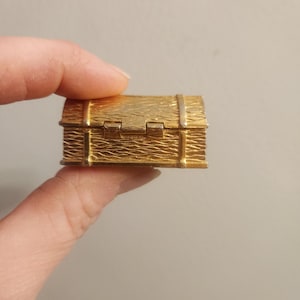 May include: A small, gold-coloured treasure chest with a textured surface. The chest has a rectangular shape with a hinged lid and metal bands. The chest is held by a person's fingers.
