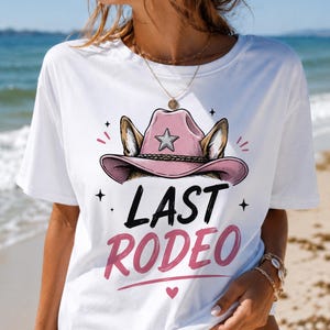 Könnte beinhalten: Weißes T-Shirt mit einem rosa Cowboyhut-Design, einem Stern und Hundeohren. Der Text "LAST RODEO" ist in Schwarz und Rosa gehalten, darunter ein kleines Herz. Ideal für einen Junggesellinnenabschied oder eine Themenfeier.