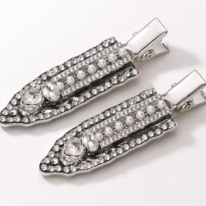 May include: Two silver-toned hair clips with a rectangular design. Each clip is embellished with clear rhinestones, faux pearls, and a heart-shaped crystal. The clips have a classic alligator style closure.