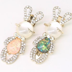 May include: Two decorative hair clips in the shape of a bunny. Each clip features a pearl head, rhinestone ears, and a rhinestone-covered body with a pear-shaped gemstone. One gemstone is pink, the other is green.