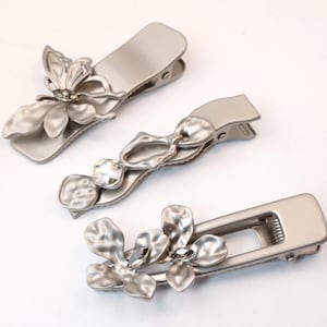 May include: Three silver-toned hair clips with floral and abstract designs. Each clip features a different decorative element, including flower and leaf shapes, and a clear crystal accent. The clips are arranged on a white surface.