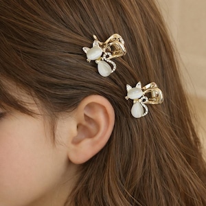 May include: Two gold-colored hair clips, each featuring a cat design with a white, oval-shaped body and sparkling accents. The clips are attached to brown hair, adding a touch of elegance and whimsy to the hairstyle. These are perfect for cat lovers.