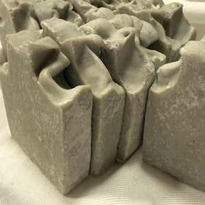 Può includere: Primo piano di diverse saponette fatte a mano. Il sapone è di colore grigio chiaro e ha una superficie strutturata. I bordi delle saponette sono irregolari, conferendo loro un aspetto rustico. Il sapone è disposto su un panno bianco.