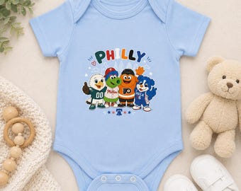 Może przedstawiać: Jasnoniebieskie body niemowlęce z nadrukiem graficznym przedstawiającym cztery maskotki sportowe z Filadelfii. Nad maskotkami znajduje się napis PHILLY kolorowymi literami. Wzór zawiera małe gwiazdki oraz ikonę dzwonu wolności.