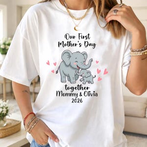 Könnte beinhalten: Weißes T-Shirt mit einer Grafik von zwei grauen Elefanten, einem großen und einem kleinen, umgeben von rosa Herzen. Der Text "Our First Mother's Day" und "together Mommy & Olivia 2026" ist auf dem Shirt aufgedruckt.