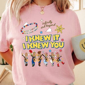 May include: A light pink t-shirt featuring a graphic print with the text "I KNEW IT I KNEW YOU" in yellow and blue. The design includes six illustrated figures holding guitars, a sheriff badge, a beaded bracelet, and the text "infinity and beyond".