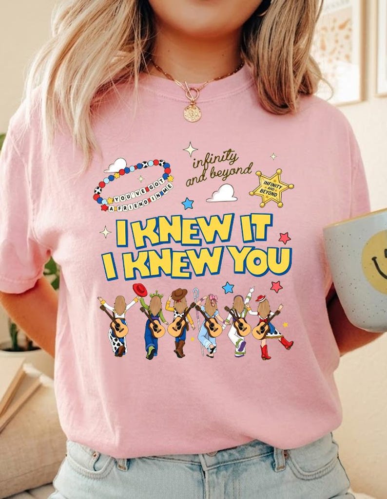 May include: A light pink t-shirt featuring a graphic print with the text "I KNEW IT I KNEW YOU" in yellow and blue. The design includes six illustrated figures holding guitars, a sheriff badge, a beaded bracelet, and the text "infinity and beyond".