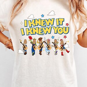 May include: A cream-colored t-shirt featuring the text "I KNEW IT I KNEW YOU" in blue and yellow. Below the text are six illustrated figures with long hair, each carrying an acoustic guitar and wearing outfits inspired by various animated characters.