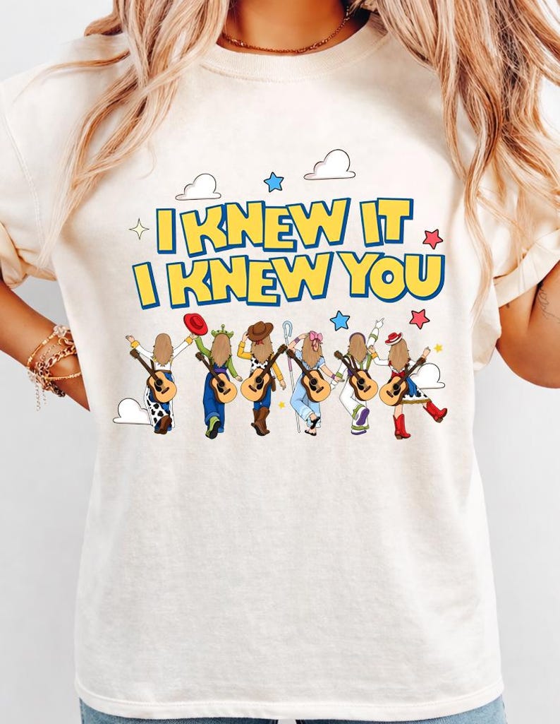 May include: A cream-colored t-shirt featuring the text "I KNEW IT I KNEW YOU" in blue and yellow. Below the text are six illustrated figures with long hair, each carrying an acoustic guitar and wearing outfits inspired by various animated characters.