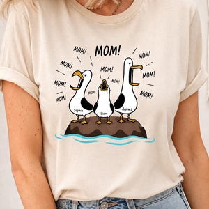 May include: Beige t-shirt featuring a cartoon illustration of three seagulls on a rock, each with a name: Sophia, Liam, and James. The seagulls are shouting "MOM!" in speech bubbles. The shirt is a casual, everyday wear item.