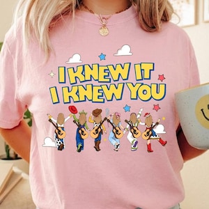 May include: A light pink t-shirt featuring a graphic print with the yellow text I KNEW IT I KNEW YOU. Below the text are six illustrated figures with long brown hair, each holding an acoustic guitar, wearing various hats and colorful outfits.