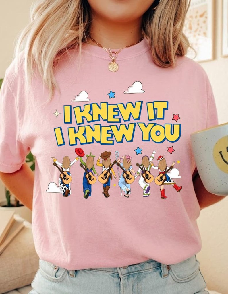 May include: A light pink t-shirt featuring a graphic print with the yellow text I KNEW IT I KNEW YOU. Below the text are six illustrated figures with long brown hair, each holding an acoustic guitar, wearing various hats and colorful outfits.