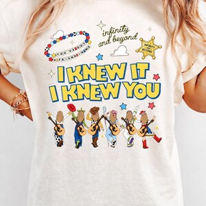 May include: A cream-colored t-shirt featuring a graphic print with the text "I KNEW IT I KNEW YOU" in blue and yellow. The design includes six illustrated figures with guitars, a beaded bracelet, a sheriff star, and "infinity and beyond" text.