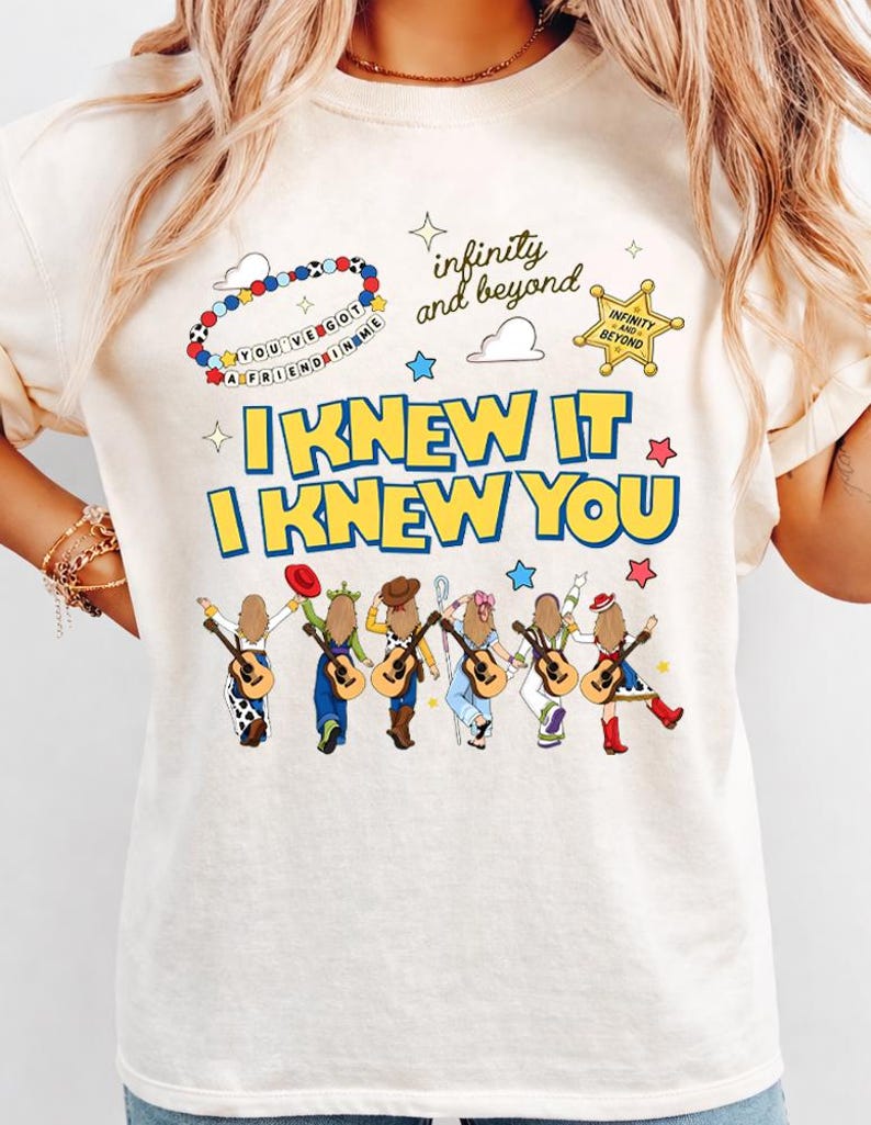 May include: A cream-colored t-shirt featuring a graphic print with the text "I KNEW IT I KNEW YOU" in blue and yellow. The design includes six illustrated figures with guitars, a beaded bracelet, a sheriff star, and "infinity and beyond" text.