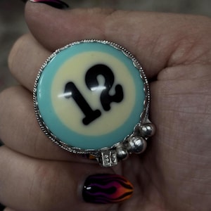 May include: A large ring featuring a pool ball design with the number 12 in black on a light blue and cream background. The ring has a silver-colored metal band and decorative accents.