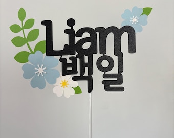 May include: A black cake topper with the name "Liam" and Korean characters. The topper is decorated with light blue and white flowers, and green leaves. It is attached to a clear plastic stick.