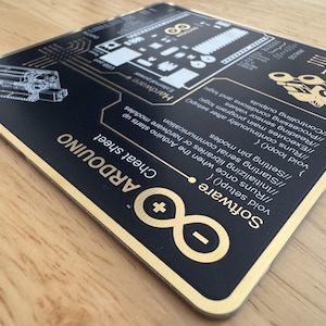 May include: A rectangular black and gold Arduino cheat sheet featuring technical diagrams, hardware labels, and software code snippets. The surface displays the Arduino logo, pin configurations, and programming syntax including void setup and void loop functions.