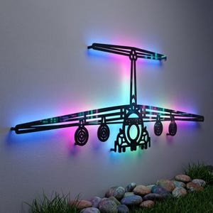 May include: A black metal wall art piece shaped like a cargo airplane silhouette. The design features a central fuselage, wings, and a T-tail stabilizer. Integrated LED lighting behind the frame emits a vibrant gradient of blue, pink, and purple.