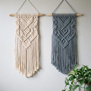 May include: Two macrame wall hangings suspended from wooden dowels. One piece is cream-coloured, and the other is slate blue. Both feature intricate knotted geometric patterns and long, straight fringe at the bottom, creating a symmetrical design.