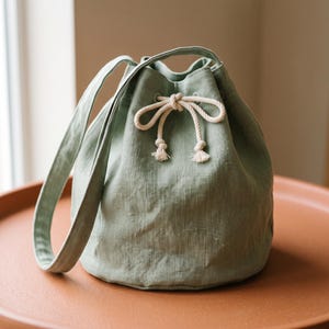 Puede incluir: Un bolso tipo saco de color verde salvia confeccionado en tela texturizada, con una correa de hombro única y un cierre de cordón ajustable asegurado con una cuerda trenzada gruesa de color blanco roto, atada en un lazo en la parte frontal.