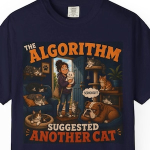 May include: A navy blue t-shirt featuring a graphic illustration of a person holding a white cat in a room filled with multiple cats and a bulldog. The text reads THE ALGORITHM SUGGESTED ANOTHER CAT and SERIOUSLY? in orange and white lettering.
