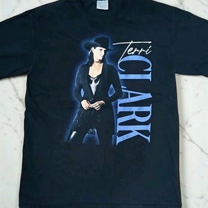 May include: Black t-shirt featuring a portrait of Terri Clark in a black hat and outfit, with the name "Terri Clark" in white script and blue block letters. The image has a blue glow effect.