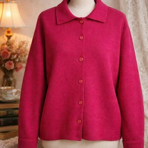 May include: A vibrant fuchsia cardigan with a classic collar and button-down front. The long-sleeved sweater is displayed on a mannequin, showcasing its tailored fit and soft texture. The bright color makes it a standout piece.