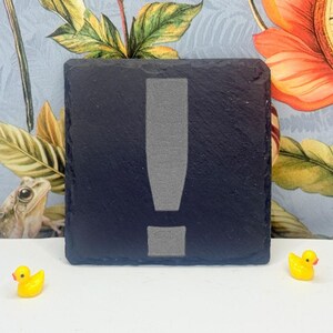 May include: A square black slate coaster featuring a centred, light grey engraved exclamation point symbol. The coaster has a rough, natural stone texture along its edges and a flat, smooth surface where the symbol is etched into the dark material.