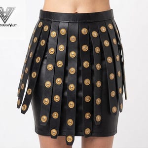 May include: Black leather mini skirt with vertical straps and gold-colored button details. The skirt has a fitted silhouette and a high waist. The design is embellished with numerous round, gold-toned buttons, adding a decorative element to the garment.