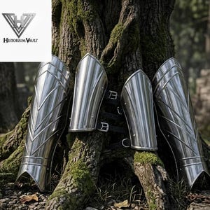 May include: A set of four polished silver metal greaves featuring a smooth, curved design with vertical ridges and black leather straps with metal buckles. The armor pieces are arranged against a textured tree trunk with a Historium Vault logo.